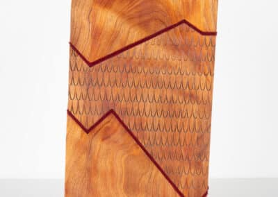 An upright wooden sculpture by Welly Fletcher featuring an abstract design. The surface has two interwoven red lines with a scale-like pattern on the top section, and natural wood grain visible on the bottom section. The sculpture is displayed on a brass base against a white background.
