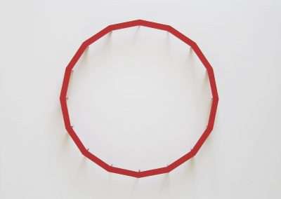 A minimalistic art piece featuring a red, irregular polygonal ring hanging against a white wall. the artwork's title and the artist's signature are at the bottom.