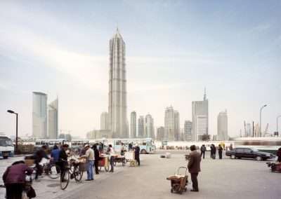 A bustling city scene captured by Horst and Daniel Zielske, featuring people and bicycles in the foreground, and a prominent, tall, needle-like skyscraper in the background amidst other modern buildings under a clear sky.