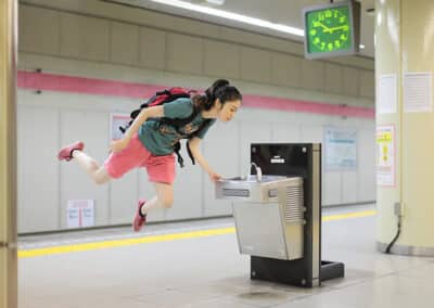 A person with a green shirt, red backpack, and red shorts appears to be floating off the ground, reminiscent of Natsumi Hayashi's whimsical photography, while drinking from a water fountain in a subway station. A green clock shows the time in the background.