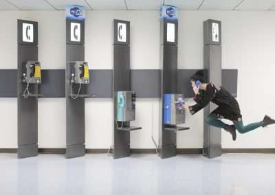 A woman, reminiscent of Natsumi Hayashi's style, playfully using a payphone, jumping mid-air with one leg bent behind her in a brightly lit indoor setting with three additional phone booths.