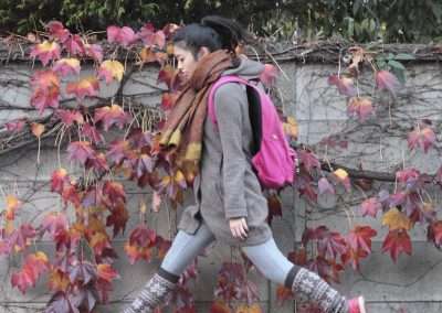 A woman in a gray coat and pink backpack walks briskly past a wall covered with red and green ivy leaves, eerily echoing the levitating spirit of Natsumi Hayashi in her dynamic stride.