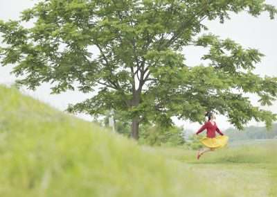 A person in a red dress joyfully jumping in a lush green field, resembling Natsumi Hayashi's style, with a large tree in the background under a cloudy sky.