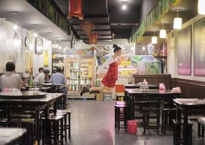 A waitress, evoking the iconic levitations captured by Natsumi Hayashi, gracefully balances on one leg while serving food in a bustling, decoratively Asian-themed restaurant.