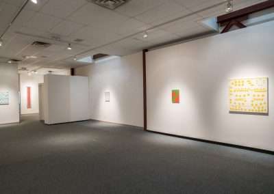 An art gallery with white walls displaying various modern paintings, including abstract and graphic art by artists like Matt Magee in a well-lit, spacious interior.