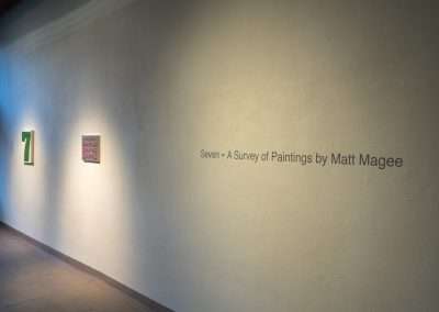 A dimly lit gallery hallway with three paintings by Matt Magee, each spaced out along a plain beige wall. Text on the wall describes the exhibit as "Seven - A Survey of Paintings by Matt Magee.