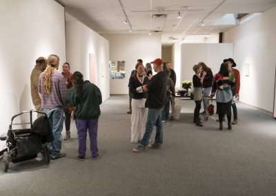 People conversing and viewing artworks at an indoor art exhibition, some standing while one person uses a mobility aid. The setting is brightly lit with white walls and pieces of art displayed.