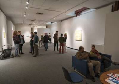 People gathered in an art gallery viewing artworks by Matt Magee on white walls, engaging in discussions. Some sit on blue chairs in relaxed postures.