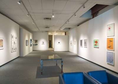 Interior of an art gallery with various camera-less framed artworks on white walls, a decorative bench at the center, and blue armchairs in the foreground. Brightly lit and spacious.