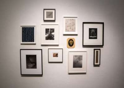 A gallery wall displaying a diverse arrangement of camera-less photographs and artwork, illuminated by soft lighting, against a white background.