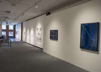 Interior of an art gallery with various camera-less framed artworks displayed on light-colored walls, including one featuring blue neon lights. A calm and quiet ambiance with minimal furnishings.