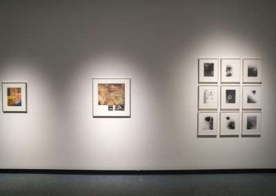 An art gallery wall showcasing three framed sets of camera-less artwork under gallery lighting, with a diverse range of styles and subjects, including both color and black and white pieces.