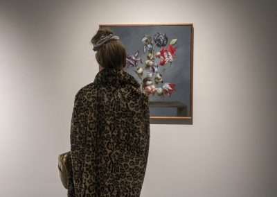 A person in a leopard print coat and a headscarf is viewing a Jason DeMarte floral painting in an art gallery.