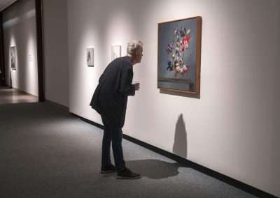 An elderly person with gray hair views a floral painting by Matthew McConville in a well-lit art gallery, standing close to analyze details, with other visitors in the background.