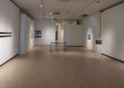 A spacious art gallery with white walls displaying various artworks, featuring a central walking path and wood flooring, dedicated to themes of spiritual journey.