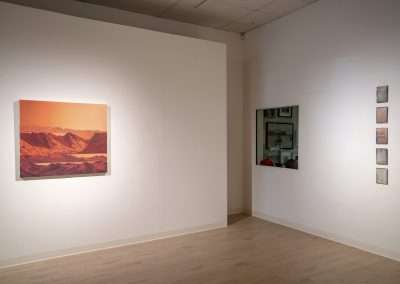 An art gallery interior with a large painting of a mountainous sunset on a white wall, symbolizing a spiritual journey, and a series of smaller framed artworks displayed sequentially on the adjacent wall.