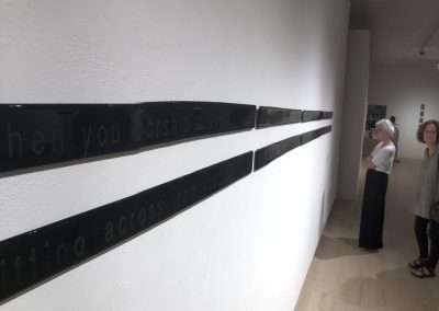 Two women observe a modern art installation featuring long black panels with white text describing pilgrimage destinations, mounted horizontally across a white gallery wall.