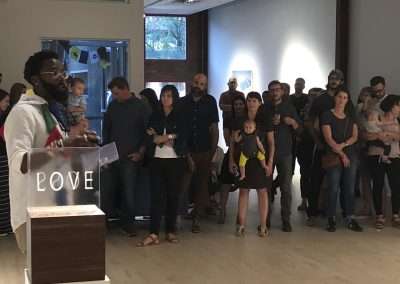 A diverse group of people stands attentively in a room listening to a speaker with glasses and a beard at a podium labeled "love." Soft lighting enhances the spiritual atmosphere.