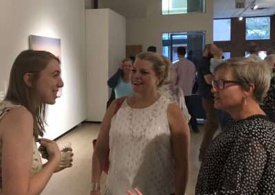 Three women chat and smile at a spiritual journey exhibition, surrounded by other attendees and artwork. One holds a drink.
