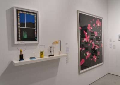 Art fair interior showing a colorful abstract painting on the left and a large graphic art piece with pink blossoms on the right, displayed beside small art objects on a white shelf.