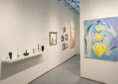 An art gallery interior featuring a white wall with various modern artworks hung neatly beside a shelf displaying artistic sculptures and objects.