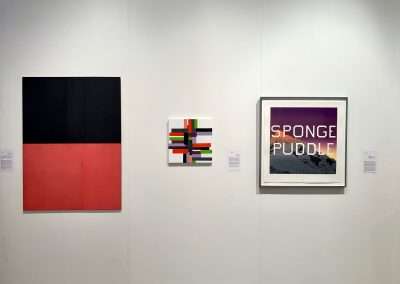 An art gallery wall displaying three framed artworks: a large abstract painting with black and red blocks on the left, a smaller colorful geometric piece in the center, and a contemporary art landscape photograph with text "sponge puddle" on the right.