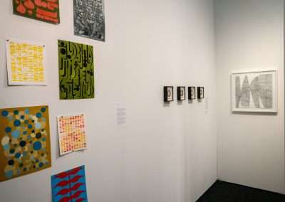 An art gallery wall displaying a variety of artworks, including abstract paintings, graphic prints with calligraphy, and small framed photographs.