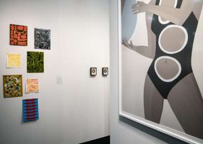 Art gallery interior featuring a large painting of a stylized figure in a black and white swimsuit, alongside several colorful abstract artworks on the surrounding walls.