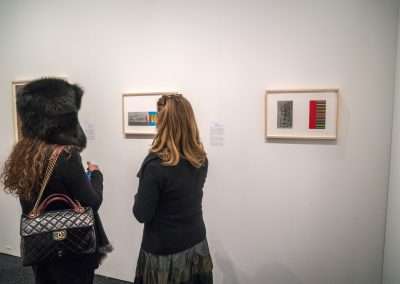 Two people viewing artwork in a gallery. one wears a large animal head mask while the other, a woman with a handbag, examines the paintings.