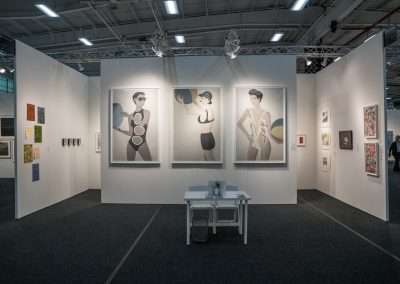 An art gallery installation with white walls featuring large portraits of stylized female figures. a table with a book and blue chairs sits centrally. artworks of various styles adorn the surrounding walls.