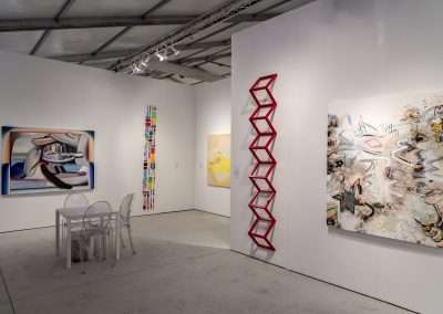An art fair interior displaying various contemporary artworks on white walls, including abstract paintings and a red sculptural piece. A table with two chairs is also present.