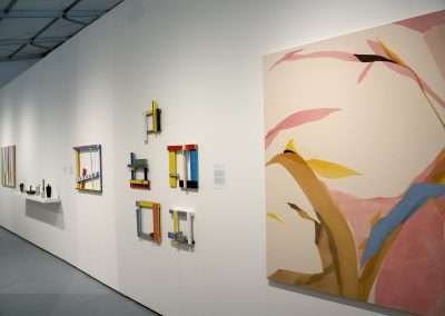 An art museum interior showing a white wall with a collection of colorful, modern abstract paintings and installations, including a large canvas with pink and yellow shapes on the right.