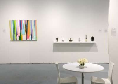 Modern art gallery interior featuring an abstract colorful painting on the left wall, a white shelf with decorative items in the center, and a white table with a bouquet of white roses.