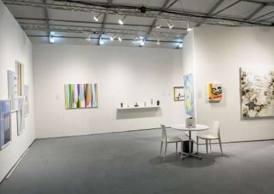 A modern art gallery with white walls, displaying various colorful paintings and small sculptures on pedestals. there's a table with two chairs in the center under bright ceiling lights.