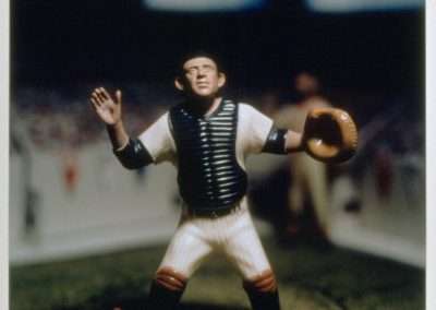 A detailed figurine of a baseball catcher in a traditional uniform, complete with a mask, chest protector, and mitt, set against a blurred stadium background.