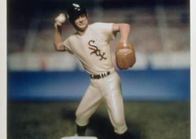 A vintage-style photograph of a miniature figurine of a baseball player in a chicago white sox uniform, posed in a batting stance on top of a base, with a blurred stadium background.
