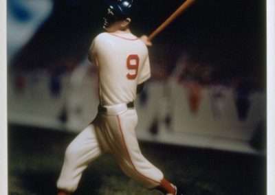 A figurine of a baseball player wearing a white uniform with the number 9, captured in a swinging pose with a bat, against a softly focused, crowd-filled stadium background.
