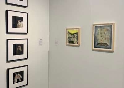 Art gallery corner displaying five framed artworks on white walls, with descriptions next to each piece. the exhibit features a mix of photography and painting.
