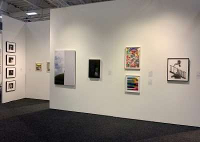 An art gallery with white walls displaying a variety of framed artworks including photographs and abstract paintings, under bright gallery lighting.