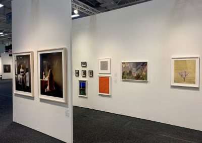 An art gallery interior showing a white wall with various framed artworks, including paintings and photographs, displayed in a well-lit exhibition space.