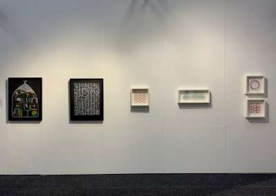 Five diverse framed artworks hung on a white gallery wall, each unique in design and color, illuminated by ceiling lights casting a soft shadow behind them.