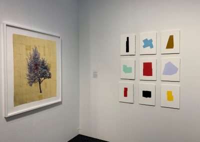 Art gallery interior displaying a wall covered with various artworks including a large image of a tree on a yellowish background and several smaller abstract colored shapes on white backgrounds.