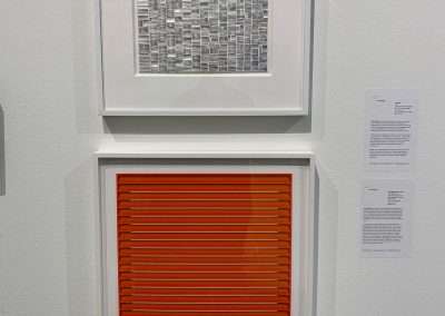 Two framed abstract artworks on a museum wall: the top piece features a grid of silver squares, and the bottom piece has horizontal orange and red stripes.