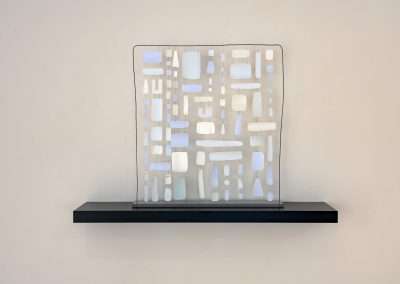 An abstract metal sculpture mounted on a black shelf against a plain wall, featuring interconnected metallic blocks and cutouts in a grid pattern.
