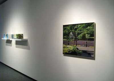 A modern art gallery interior featuring paintings and a book display on white walls under directed lighting, with a focus on a prominent painting of a verdant, abstract landscape.