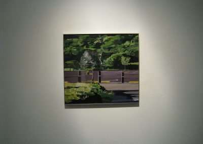 A painting of a green garden scene with a fence and foliage hangs on a white gallery wall, illuminated by a spotlight.