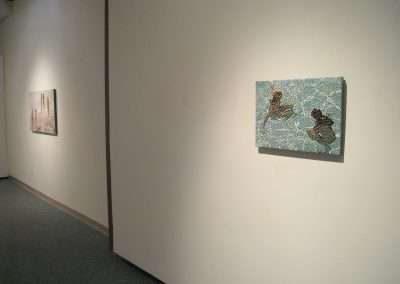 A bright art gallery interior featuring two paintings mounted on a white wall, with one painting on the left showing a landscape and another on the right with abstract design.