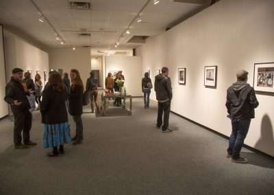 People viewing and discussing photographs at an art gallery exhibition with framed pictures on white walls, and refreshments available on a central table.