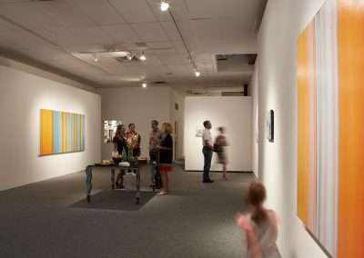 An art gallery scene with visitors viewing colorful, abstract paintings. a group gathers by a table with refreshments, and a child blurs in motion in the foreground.