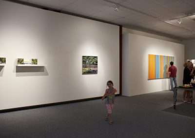 An art gallery room with white walls displaying various framed photographs. a young girl walks towards the camera, and two adults observe artworks in the background.
